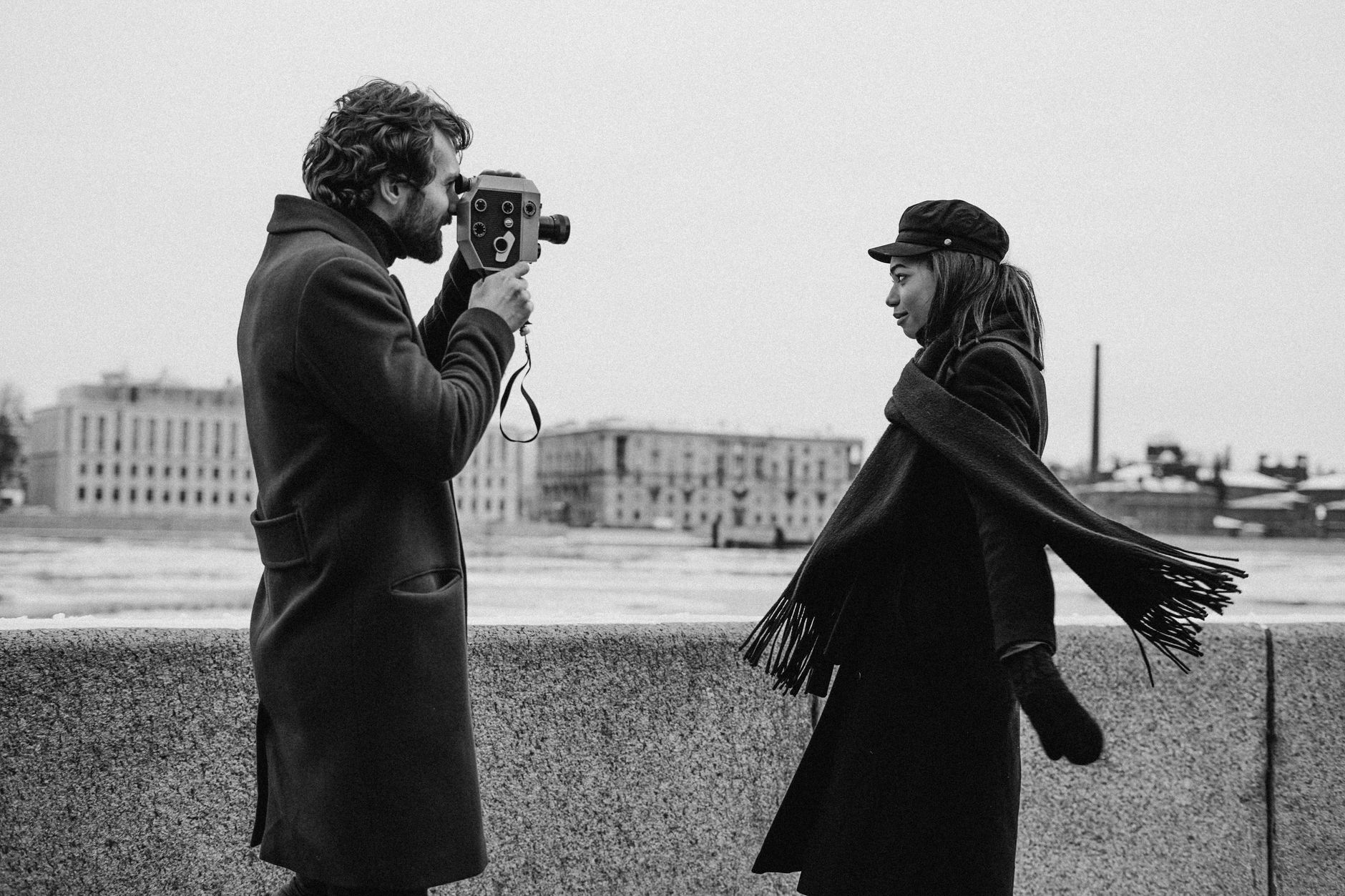 A couple in winter attire poses with a vintage camera in a black and white photoshoot.